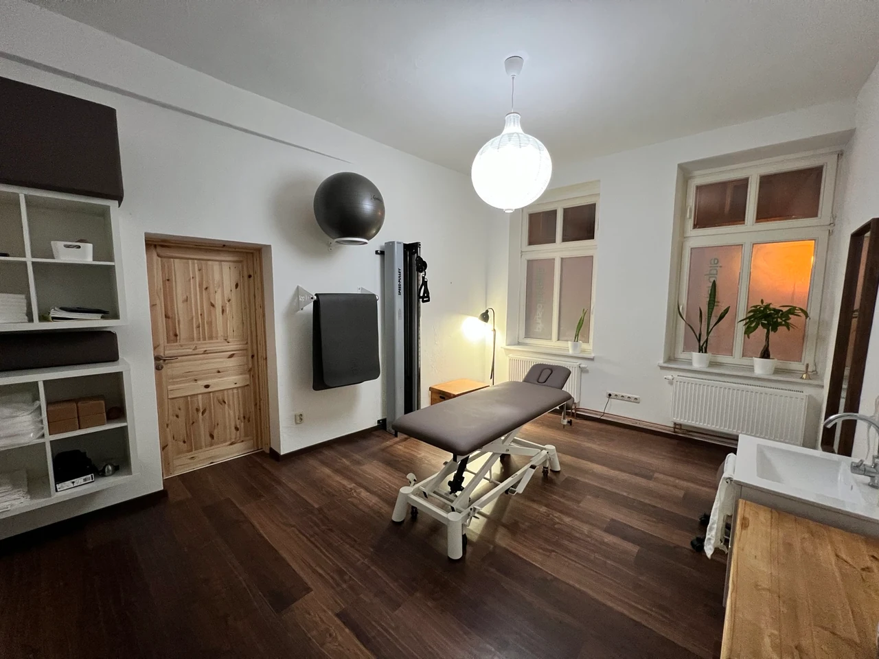 Behandlungsraum der Physiotherapie am Wall mit Behandlungsliege, Trainingsgerät, Holzfußboden und Fenstern
